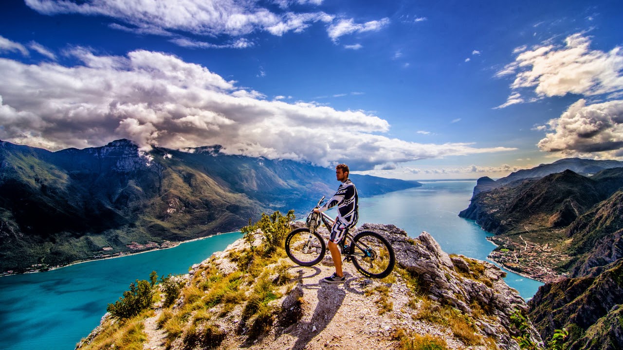 Mountain biker with a bicycle on a rocky overlook facing a turquoise lake surrounded by green mountains under a cloudy sky